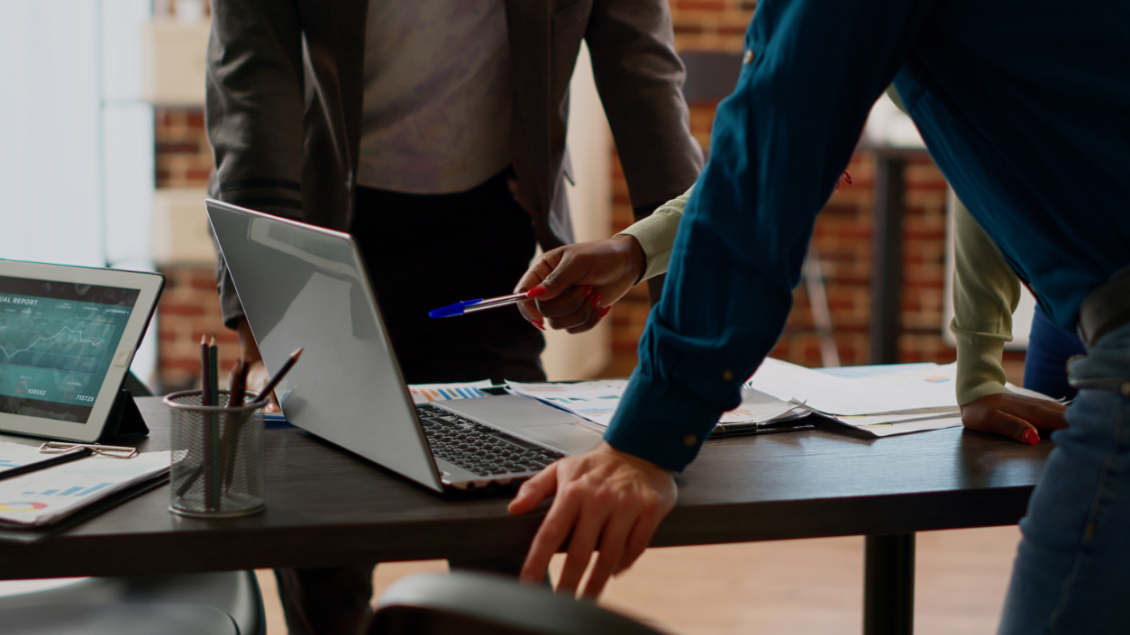 Colleagues lean over a desk to review a laptop together, and one of them points at the screen with a pen. On the desk are a tablet showing an annual report, printed charts and a pen pot.